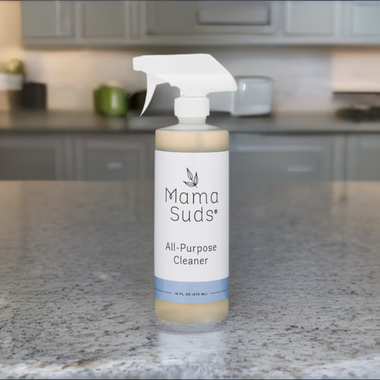 A bottle of MamaSuds All-Purpose Cleaner Spray, made with natural ingredients, rests on a granite countertop in a modern kitchen with blurred cabinets in the background.