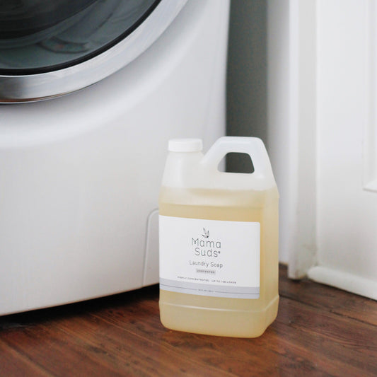 A container of MamaSuds Laundry Detergent Soap, made with natural ingredients, sits on a wooden floor next to a white washing machine.