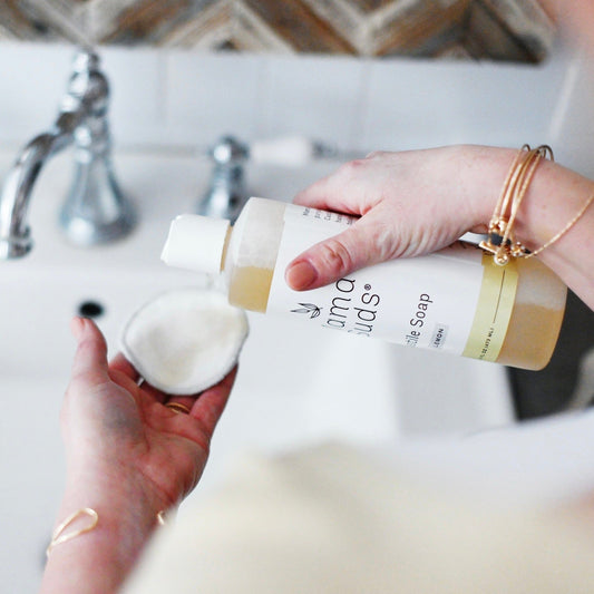 A person pours MamaSuds Castile Liquid Soap from a bottle onto a round sponge over a sink, wearing gold bracelets. A silver faucet and tiled wall are visible in the background.