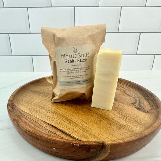 Two biodegradable, vegan MamaSuds Laundry Stain Sticks—one wrapped, one unwrapped—sit on a round wooden tray with a white subway tile background.
