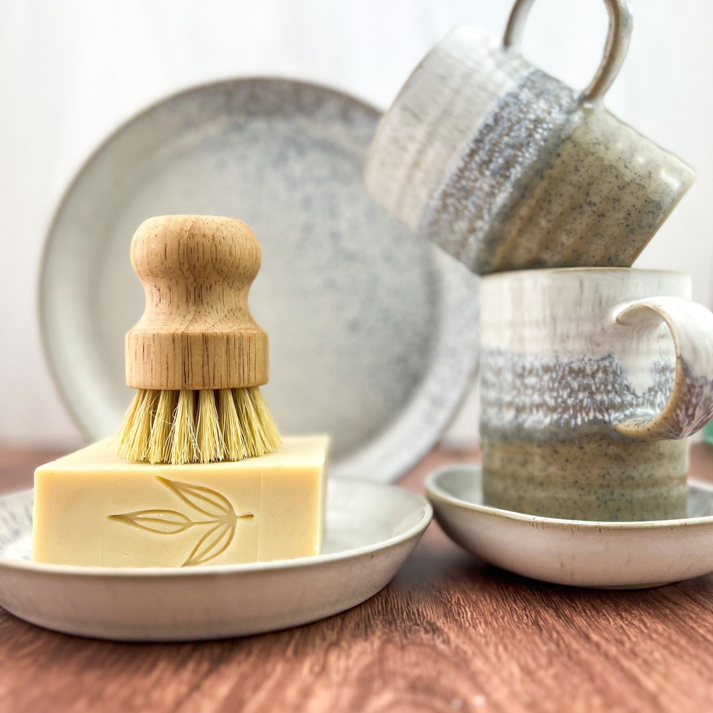 A wooden-handled scrub brush sits atop MamaSuds Solid Dish Soap with a leaf design on a ceramic plate. Two speckled mugs stack on matching plates behind, all on wood. This vegan, biodegradable soap cleans sustainably.