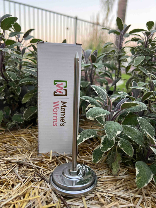 A Memes Worms metal thermometer stands in straw among green plants on a worm farm, near a box labeled Memes Worms with a colorful logo; a fence and palm tree appear blurred in the background.