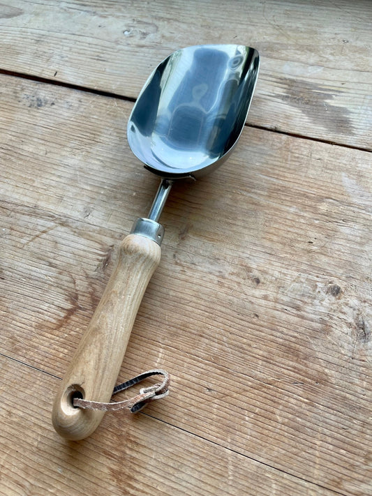 A metal garden scoop with a wooden handle and leather strap from The Celtic Farms Potters Tool Set - 3 Tools for Potting rests on rustic wood, making it a perfect Garden Tool Gift or addition to any potting toolkit.