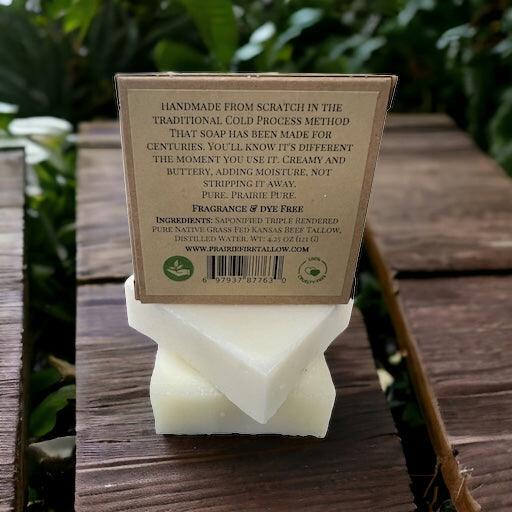 A Pure Beef Tallow Soap Bar by Prairie Fire Tallow, Candles, and Lavender sits on a wooden surface, its label noting the traditional cold process and natural organic ingredients. Lush greenery is softly blurred in the background.