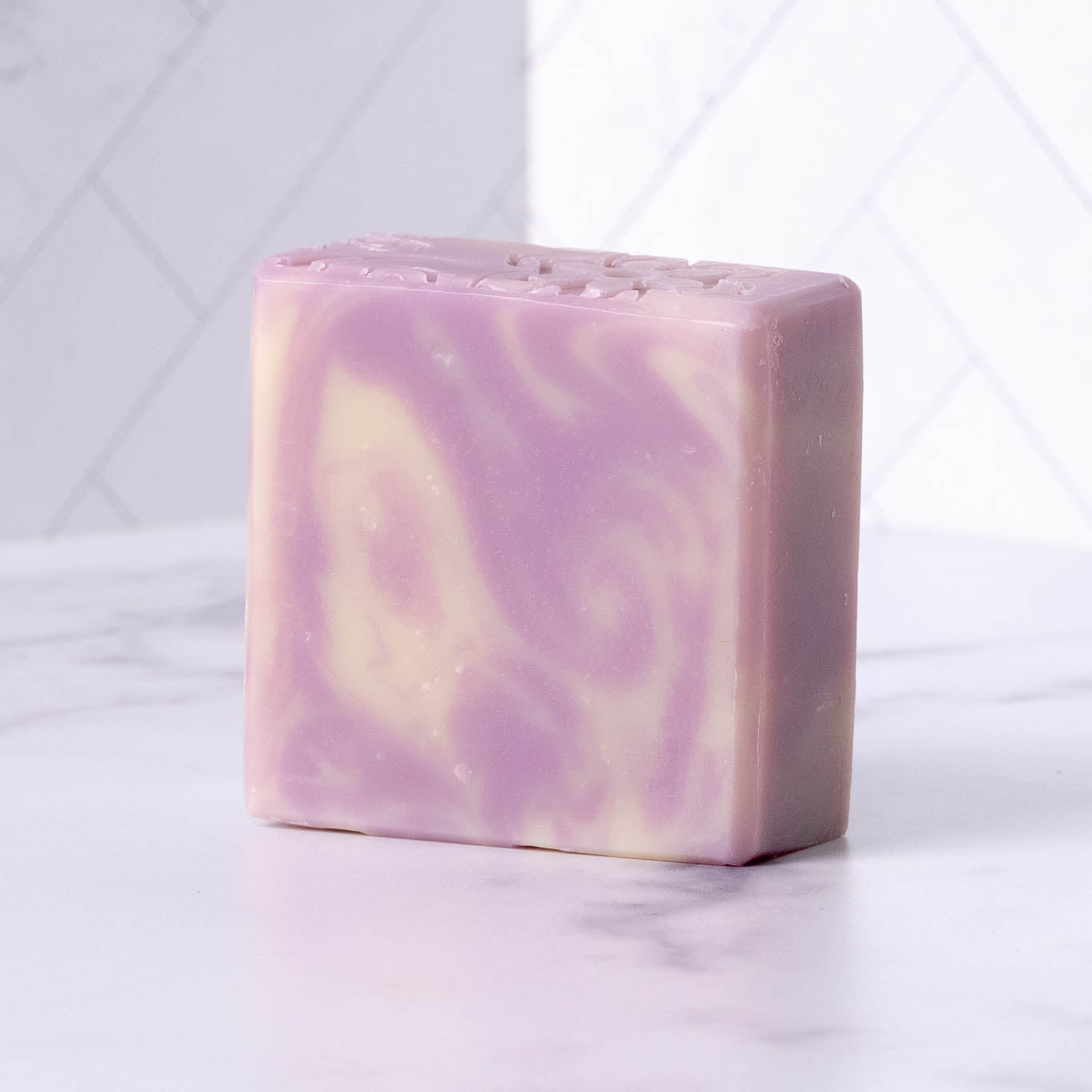 Rustic Strength Bar Soap | Lavender, a square bar with a marbled cream pattern, sits on white marble. Perfect for sensitive skin, this soap is shown against a backdrop of white tiles.