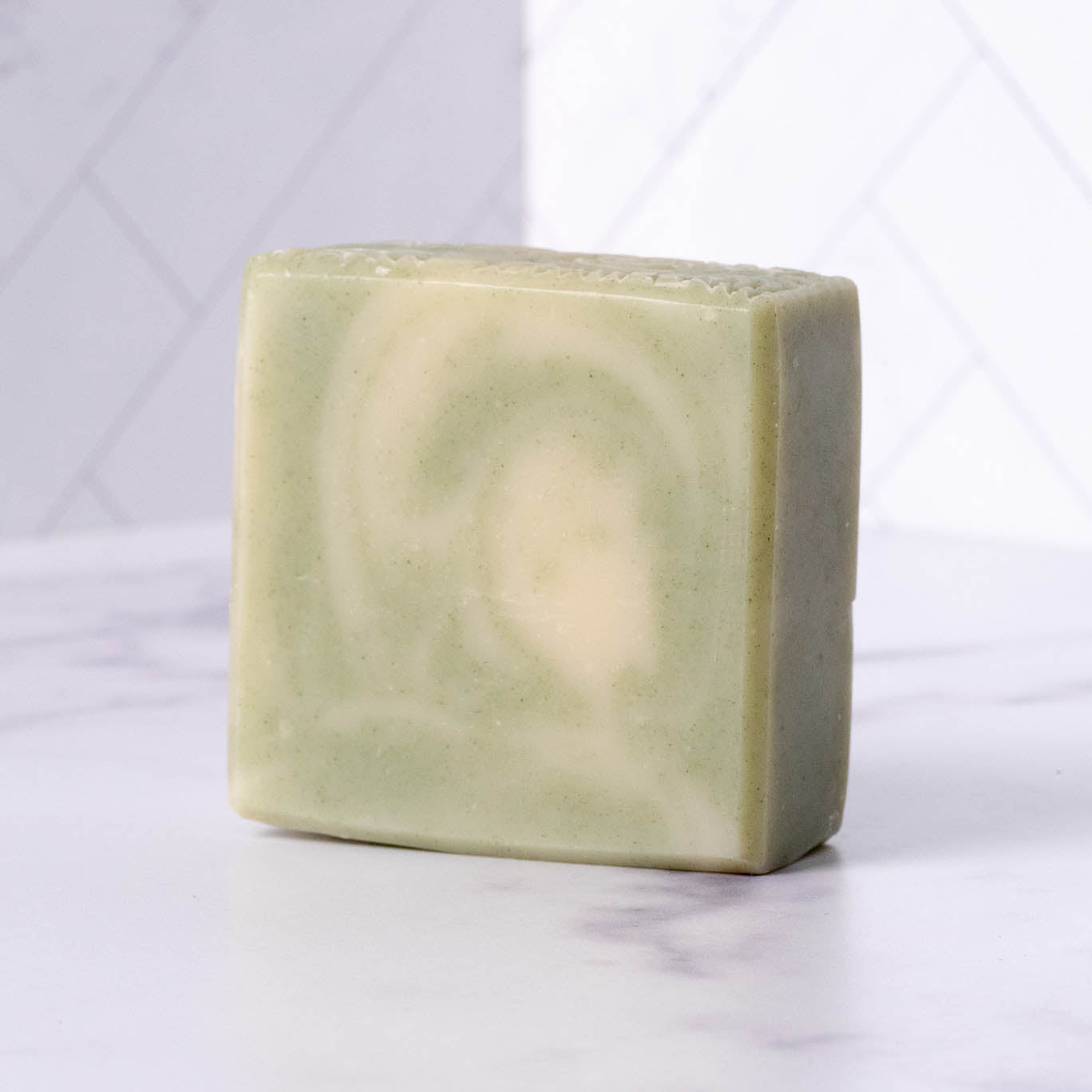 A rectangular bar of Rustic Strength Bar Soap | Peppermint Tea Tree, pale green in color with a subtle abstract pattern, rests on a white marble surface against a white tiled wall.