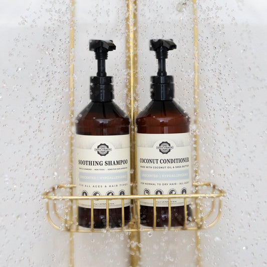 Two brown pump bottles—one labeled Soothing Shampoo and the other Rustic Strength Coconut Conditioner | Mama + Baby Safe—rest on a gold shower caddy, surrounded by water droplets inside a shower.
