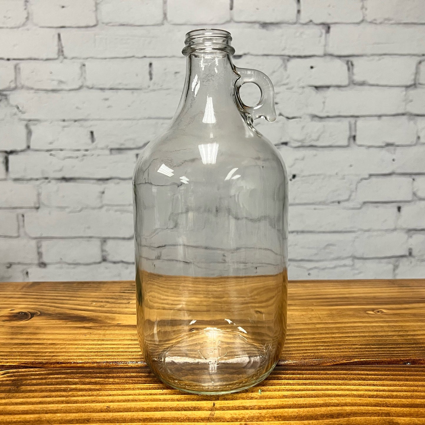 The Rustic Strength Half Gallon Glass Jug with Cap, featuring a small handle near the neck, sits on a wooden surface in front of a white brick wall—perfect for your needs and available in limited stock.
