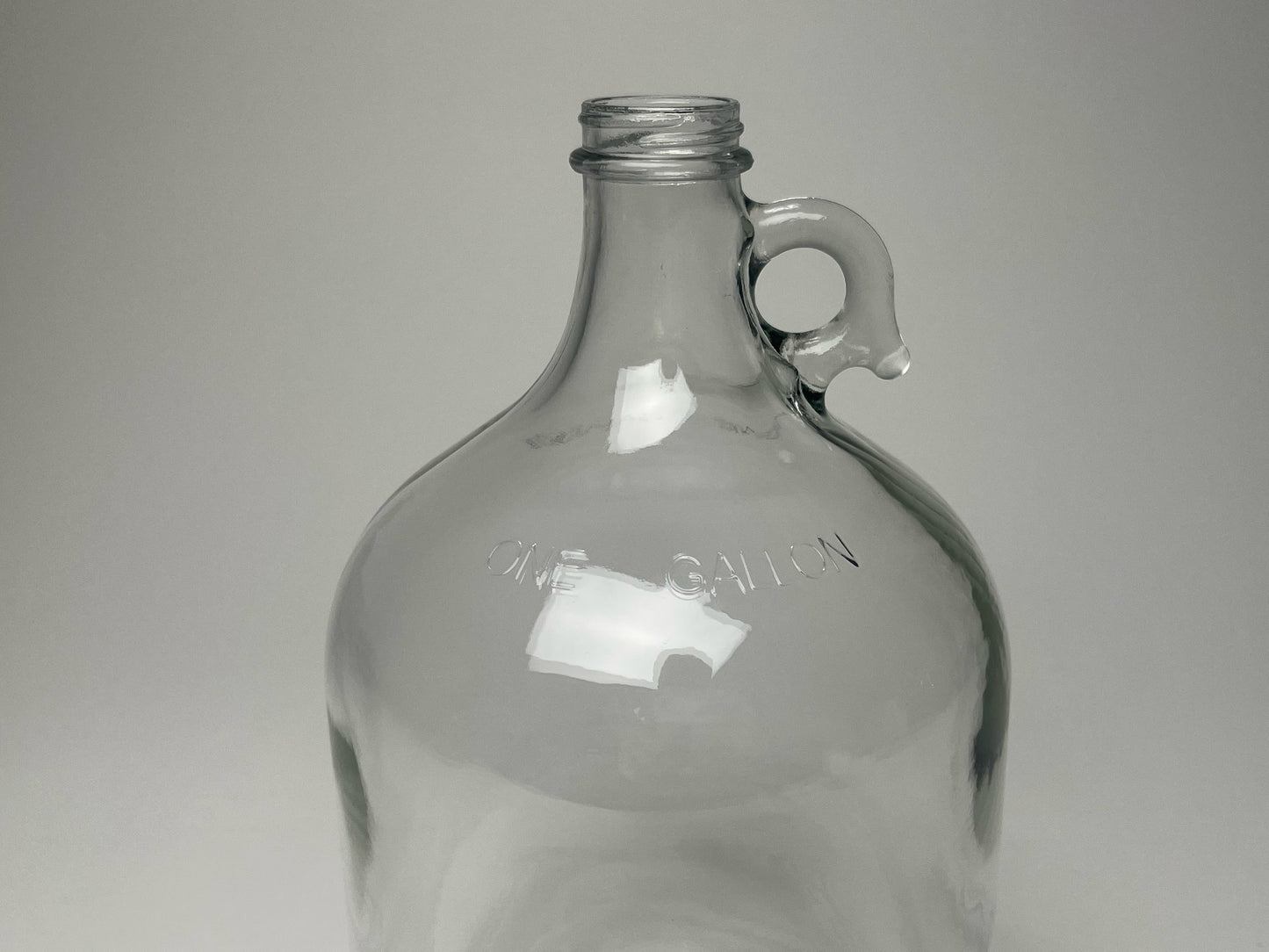 The Rustic Strength 1 Gallon Glass Jug with Cap features a small handle near the neck and ONE GALLON embossed on the shoulder. Displayed against a light gray background. Limited stock available.