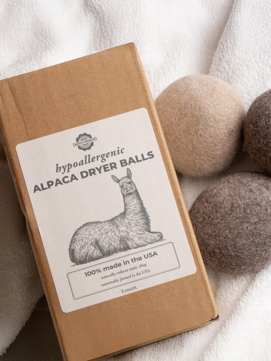 A box of Rustic Strength Alpaca Dryer Balls (3 pack), featuring an alpaca illustration, sits beside three natural-shaded wool alternative balls on soft white fabric for gentle static reduction.