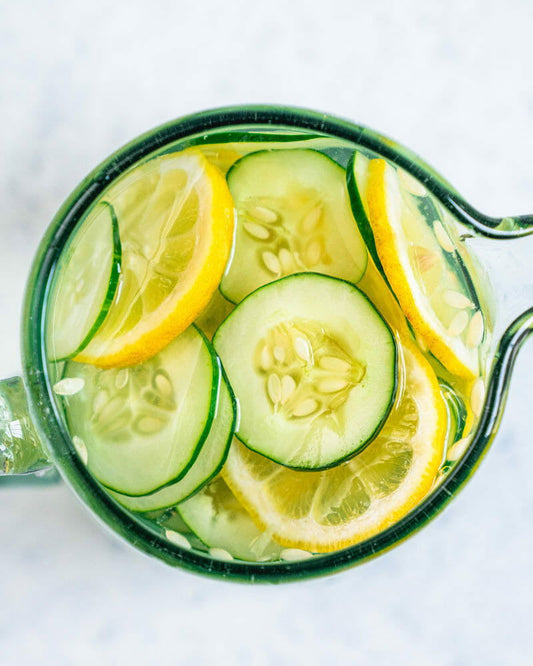 A glass pitcher with water, cucumbers, and lemon slices rests on a marble surface, evoking the calming essence of Salacia Salts' Midnight Garden Small Salt Soak.