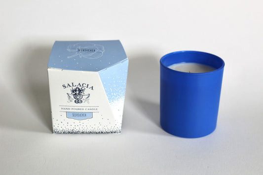 A blue hand-poured soy blend Seersucker Signature Candle by Salacia Salts rests in a matching glass holder beside its white box with blue accents and an elegant illustration.