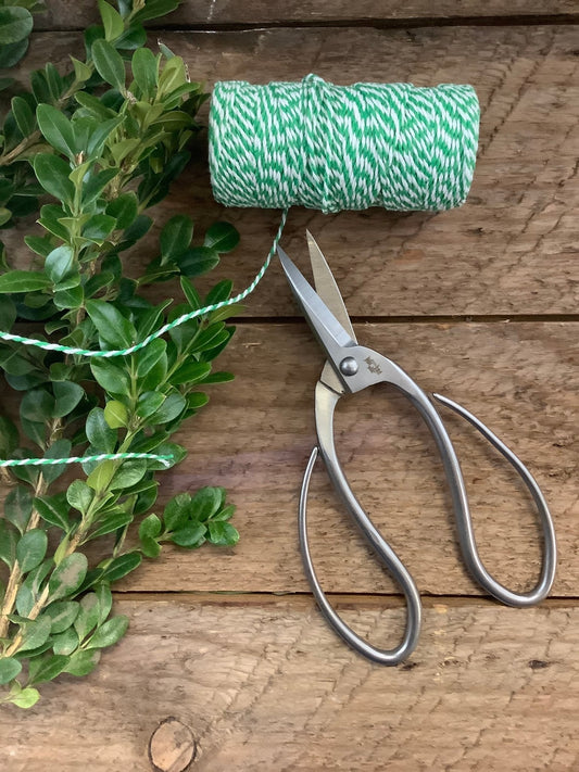 The Celtic Farm Stainless Garden Scissors rest on a rustic wooden surface beside green and white twine, which is partially cut and attached to the scissors, along with green leafy stems.