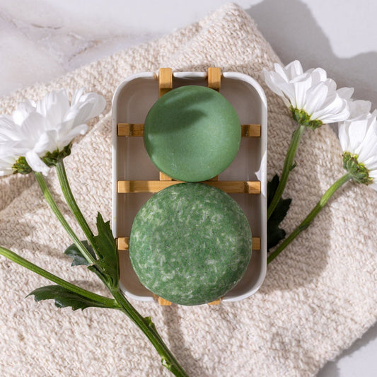 Two green Bloom Castor Oil Conditioner Bars by ZeroWasteStore.com rest on a wooden soap dish atop a beige towel with daisies, offering plastic-free, nourishing care for stronger, softer, healthier hair.