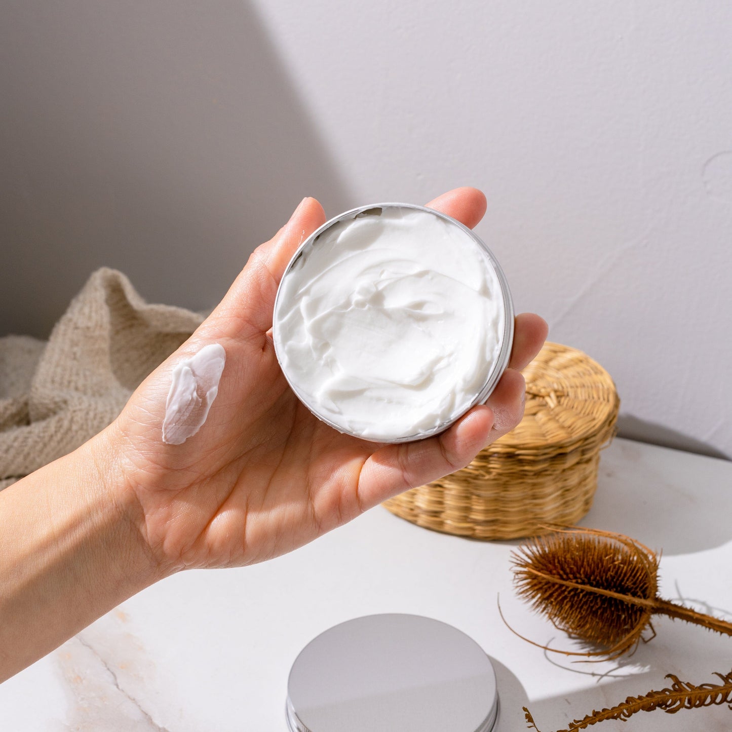 A hand holds an open tin of ZeroWasteStore.com’s Zero Waste Body Butter - Shampoo Bar Scents (4oz), deeply nourishing and moisturizing, with a dab on the skin. A basket, brush, and beige fabric rest on a white surface in the background.