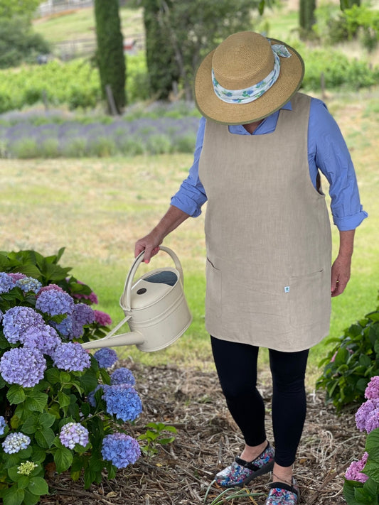 Wearing a straw hat, blue shirt, leggings, and The Celtic Farm Linen Apron - French Style Crossback, a person waters blooming hydrangeas in a lush garden.