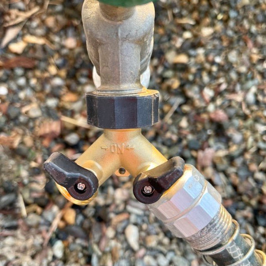 Close-up of The Celtic Farm Two-way Brass Garden Hose Splitter, showing its leak-proof brass spigot and Y-shaped design that connects two hoses. Visible valves sit among small rocks and mulch in the background.