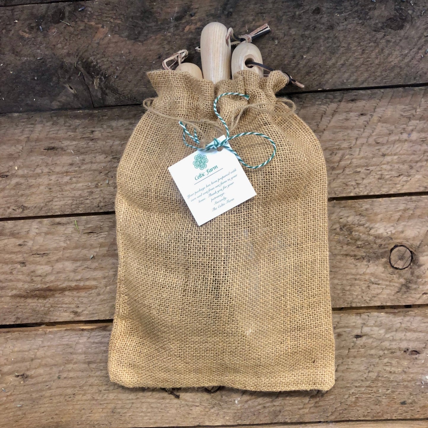 A burlap sack with a twine-wrapped tag rests on rustic wood, partially revealing The Celtic Farm’s Unique Garden Tool Set & Gift—quality-crafted garden tools with wooden handles inside.