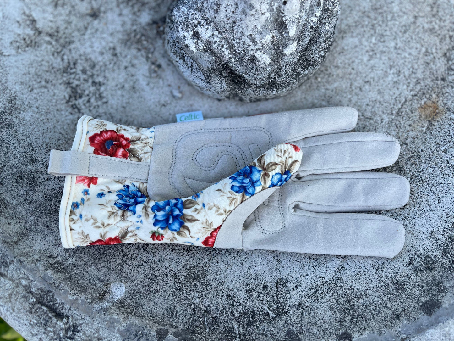 A pair of The Caroline Womens Floral Gardening and Project Gloves by The Celtic Farm, with a blue and red floral cuff, rests on a weathered stone surface next to a round stone ornament.