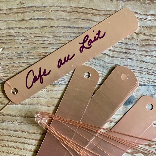 The Celtic Farms Copper Garden Tags with Wire (5 inches), labeled Cafe au Lait in cursive and featuring an aged patina, rest on wood beside blank tags and copper wires—perfect as a thoughtful gift for gardeners.