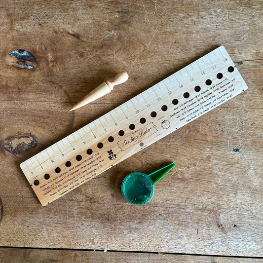 The Celtic Farm Seeding Gift Set - Dibble, Seeder, and Ruler includes a wooden ruler with measurements and planting holes, a wooden dibber, and a small green container—perfect gardening tools arranged on a wooden surface.