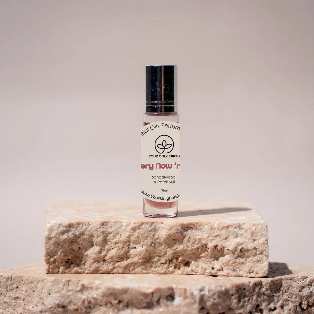 A small glass bottle of Perfume Roll-on from Your Only Earth, labeled Sandalwood & Patchouli and blended with organic fractionated coconut oil, sits on two stacked stone blocks against a neutral backdrop. It features a black cap and simple, elegant branding.