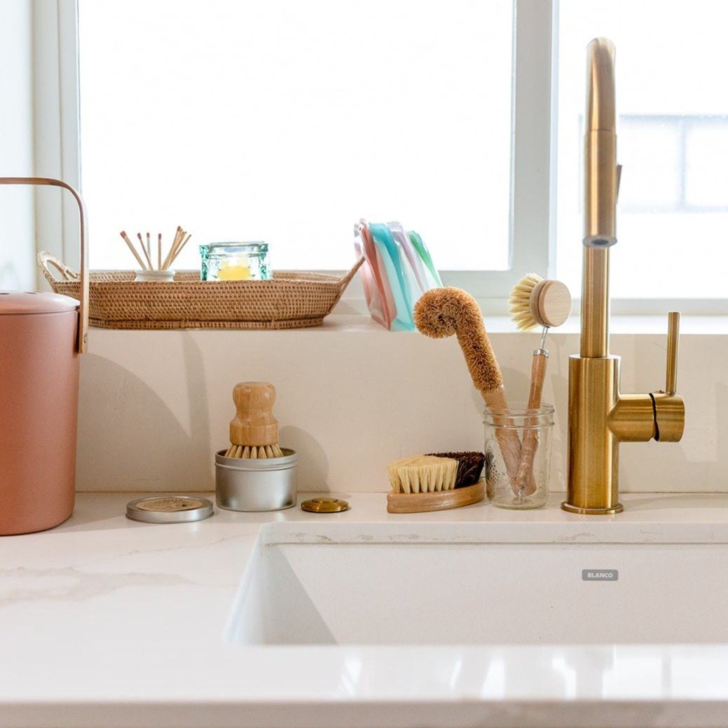 On a white countertop by a window, a gold faucet and modern sink feature ZeroWasteStore.com’s Bottle Cleaning Brush in jars, with eco-friendly brushes, a pink compost bin, colorful dish towels on a wicker tray, and small items.