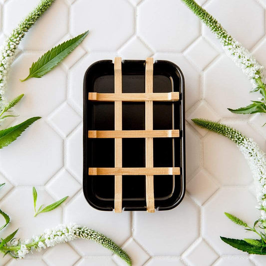 The Bamboo Soap Dish by ZeroWasteStore.com, featuring a black rectangular design with a wooden lattice cover, rests on white hexagonal tiles accented by green leaves and white flowering stems.