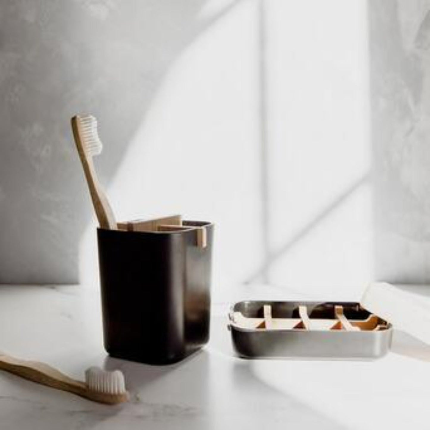 The Zero Waste Brush Holder & Soap Dish Bundle from ZeroWasteStore.com features bamboo toothbrushes in a black holder, a compostable brush on a white surface, and an open stainless steel soap dish—sunlit shadows create an eco-friendly bathroom scene.