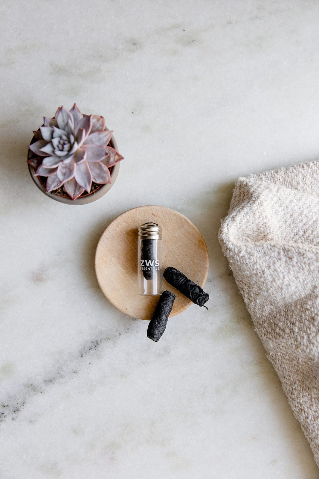 A small potted succulent, a wooden dish with a ZeroWasteStore.com glass bottle of Bamboo Charcoal Floss, and two black floss refills rest on marble beside a textured beige towel.