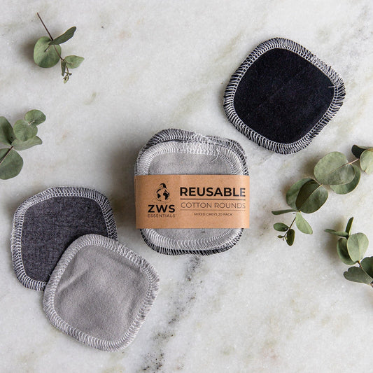 Four reusable cotton rounds from ZeroWasteStore.com, in black and grey, are displayed on marble with eucalyptus sprigs. These eco-friendly rounds come packaged with a brown label reading "Reusable Cotton Rounds.