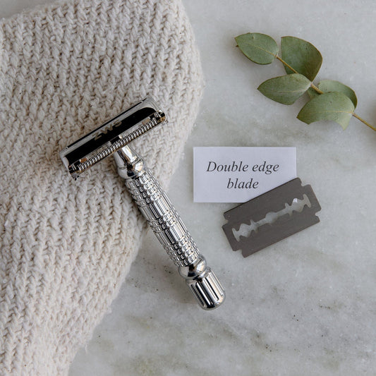 The ZeroWasteStore.com Zero Waste Safety Razor—unisex, single blade, with 10 free blades—rests on a white cloth with a razor blade, note saying Double edge blade, and eucalyptus sprig atop marble for plastic-free grooming.