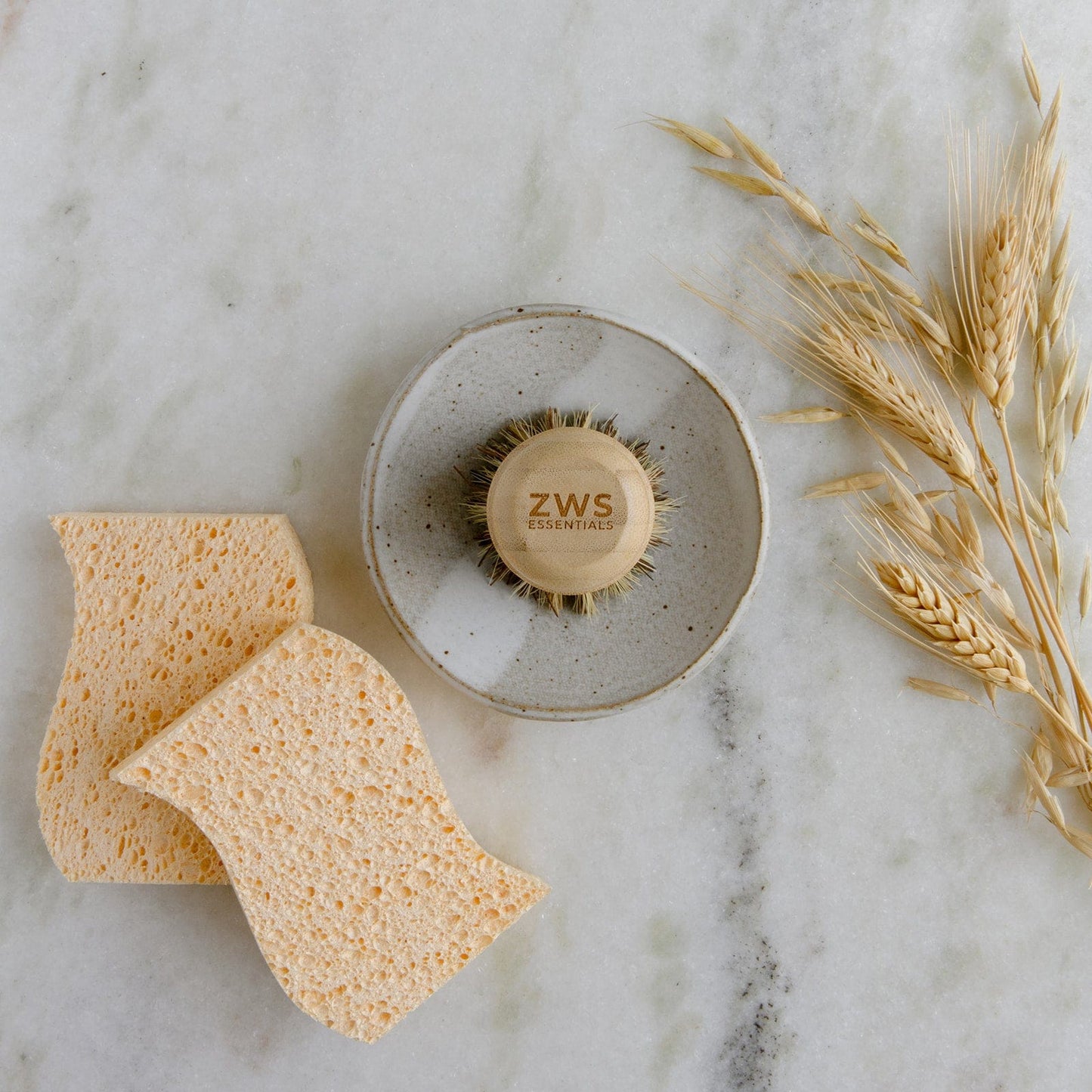 The ZeroWasteStore.com Dish Sponge Mini Kit, featuring a round plant-based brush in a ceramic holder and two zero-waste sponges, is displayed on a white marble surface alongside wheat stalks.