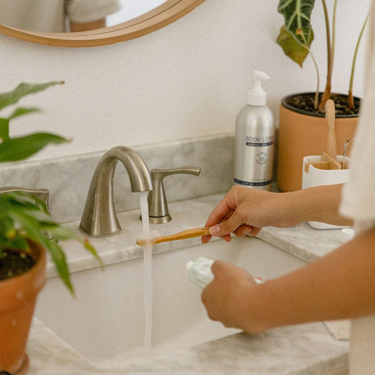 A person holds a Bamboo Toothbrush from ZeroWasteStore.com under running water in a bathroom sink, ready to use. Plants and a body lotion bottle decorate the marble countertop.