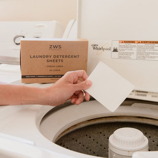 A hand holds a ZeroWasteStore.com Laundry Detergent Sheet over an open washing machine, with the eco-friendly detergent sheets box and Whirlpool machine lid visible in the background.