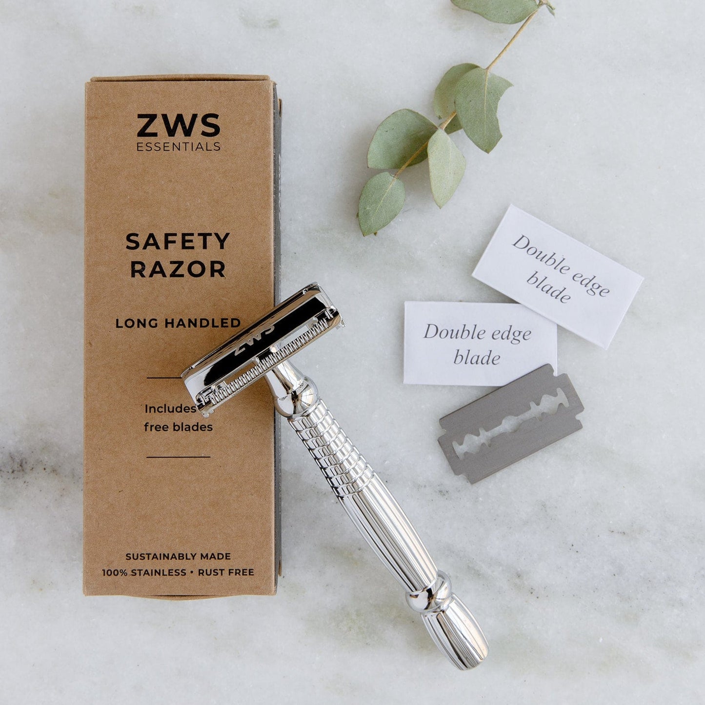 A ZeroWasteStore.com Zero Waste Safety Razor with a single blade, 10 free blades, and a brown box lies beside green eucalyptus leaves on white marble—a sustainable, plastic-free unisex razor for smooth shaves.