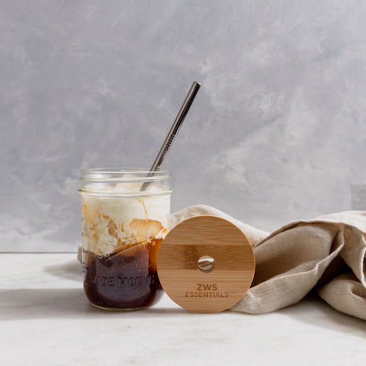 A Mason Jar Kit from ZeroWasteStore.com—with a glass mason jar, bamboo lid, and stainless steel straw—holds iced coffee and cream, while the branded lid rests on a beige cloth against a light gray background.