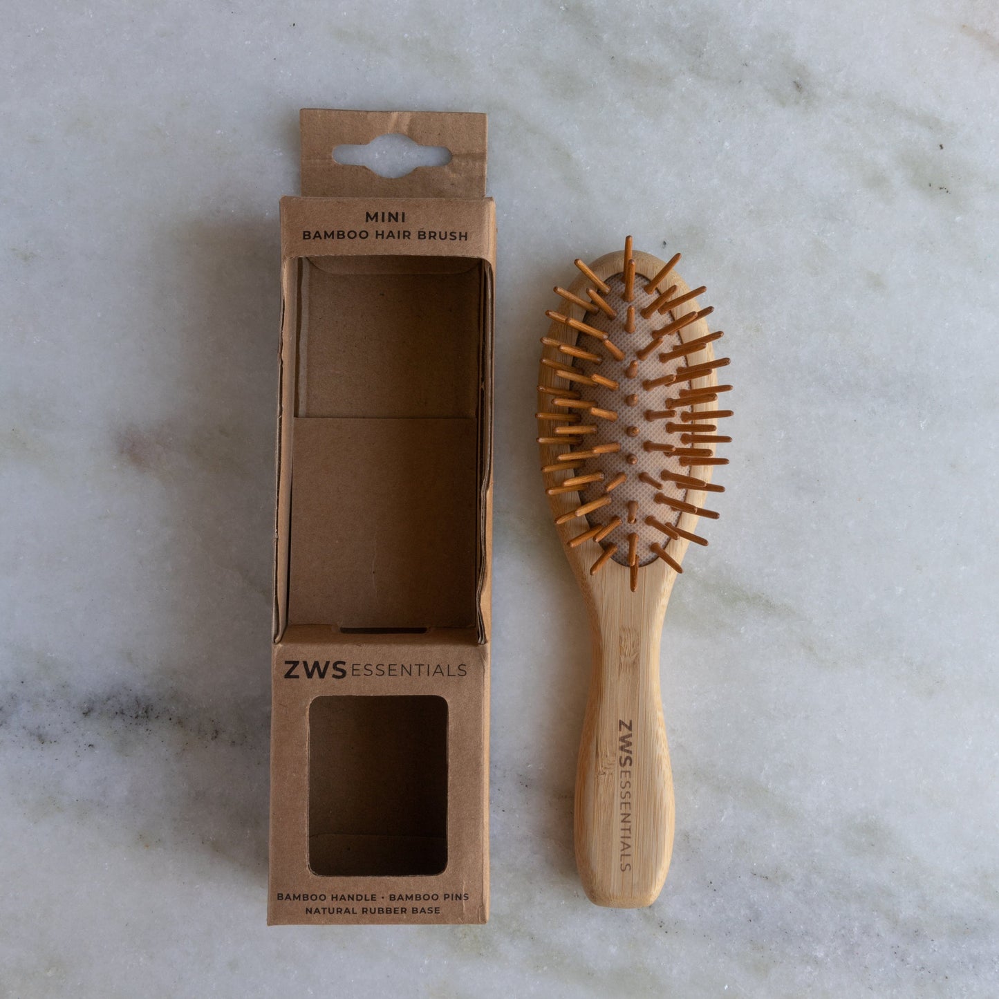 The ZeroWasteStore.com Mini Bamboo Hair Brush—100% bamboo, plastic free, compostable—rests beside its open cardboard box labeled “Mini Bamboo Hair Brush” on a light marble surface.