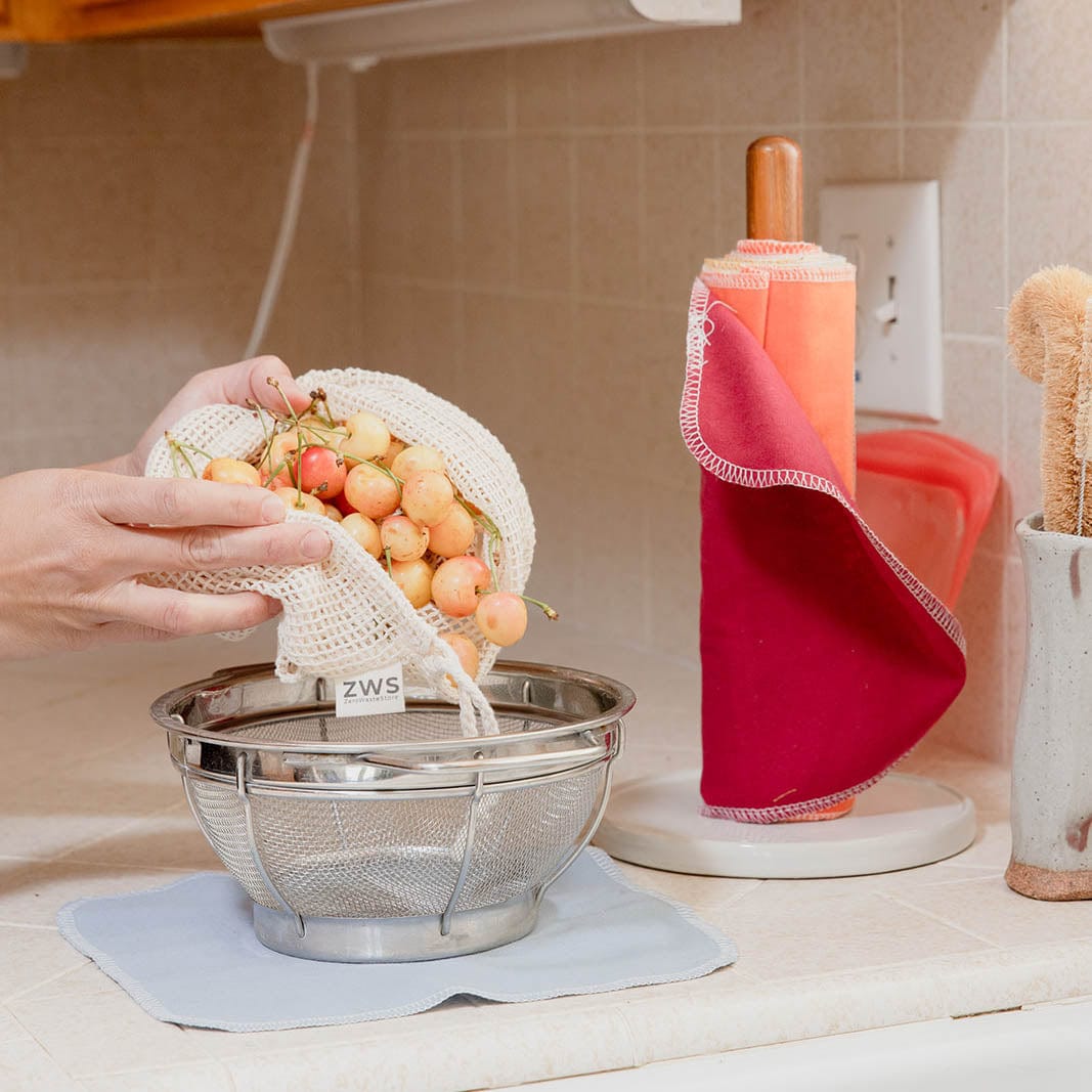 Someone pours light-colored cherries from a ZeroWasteStore.com Mesh Produce Bag - Multiple Sizes - Zero Waste Net Bag into a metal strainer on the kitchen counter, surrounded by cloth towels and a dish brush for a zero waste lifestyle.