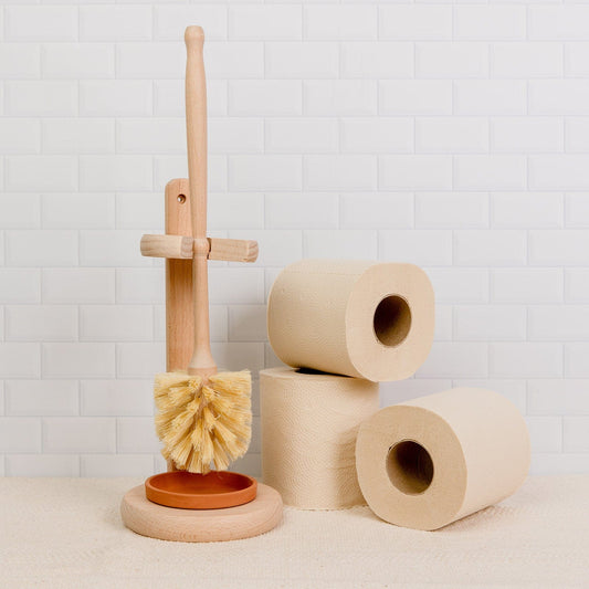 A Plastic-Free Toilet Brush by ZeroWasteStore.com with its stand is placed beside three beige toilet paper rolls against a white tiled wall.