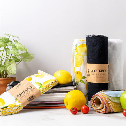 ZeroWasteStore.com Reusable Paper Towels, made with organic cotton and featuring bright patterns and lemon prints, are stacked beside fresh lemons, tomatoes, a green apple, and a small potted plant on a white surface.