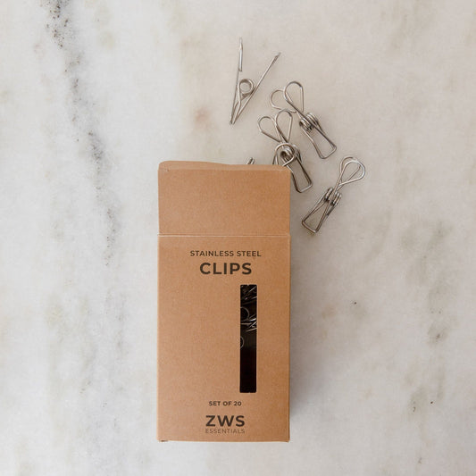 A brown cardboard box labeled "Stainless Steel Clips - Laundry Pegs, Clothespins, Multipurpose Clips - 20 Pack" by ZeroWasteStore.com sits on white marble, with four metal clips visible—ideal for a sustainable, plastic-free laundry routine.