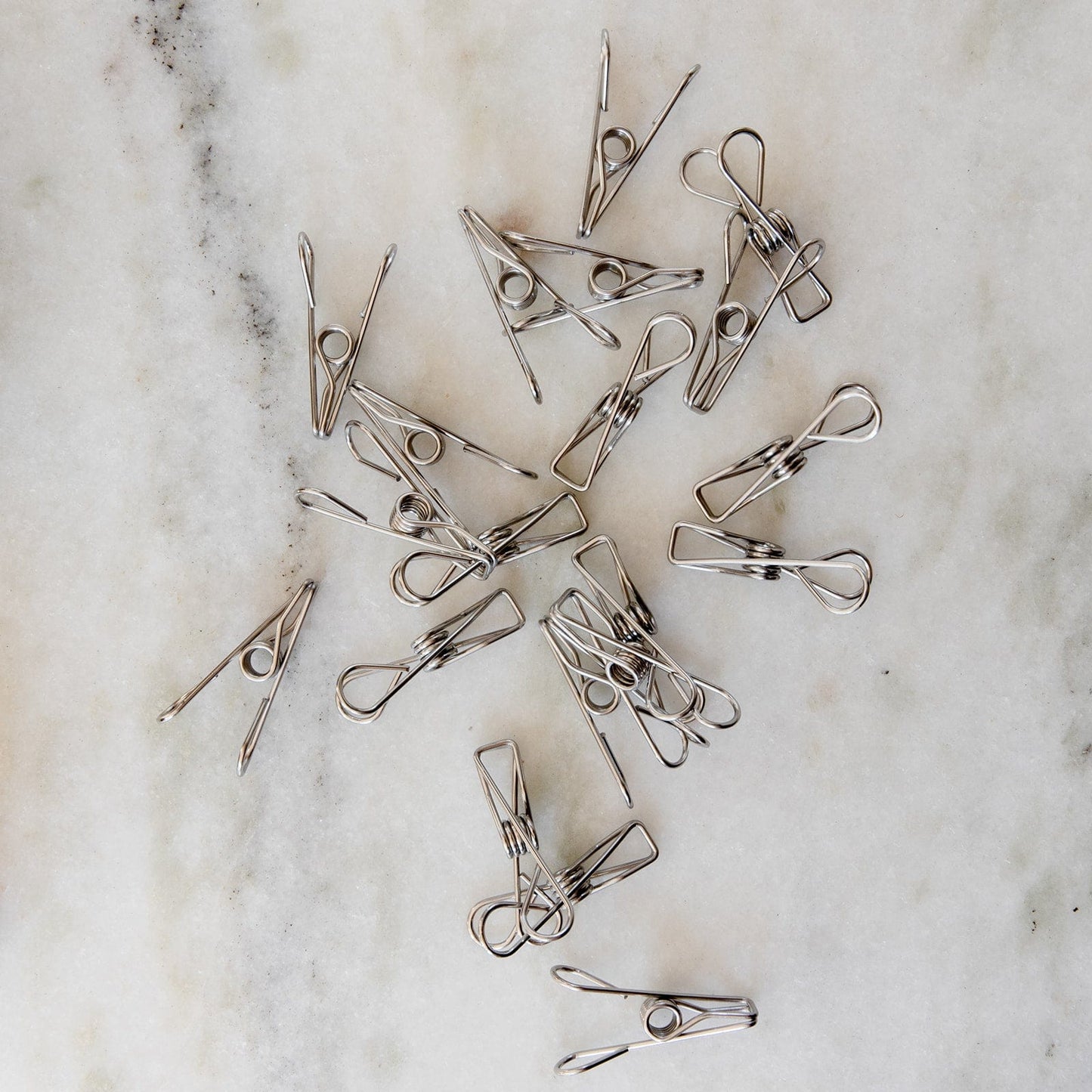 ZeroWasteStore.com Stainless Steel Clips—Laundry Pegs, Clothespins, Multipurpose Clips (20 Pack)—are scattered on a marble surface, showcasing their simple, eco-friendly design as a stylish plastic-free alternative for everyday use.