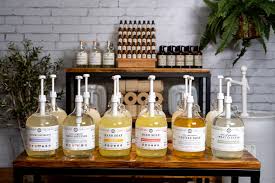 A display of large refill bottles with pumps, containing various liquid soaps or cleaners, set on a wooden table with smaller glass bottles, plants, and paper rolls in the background.