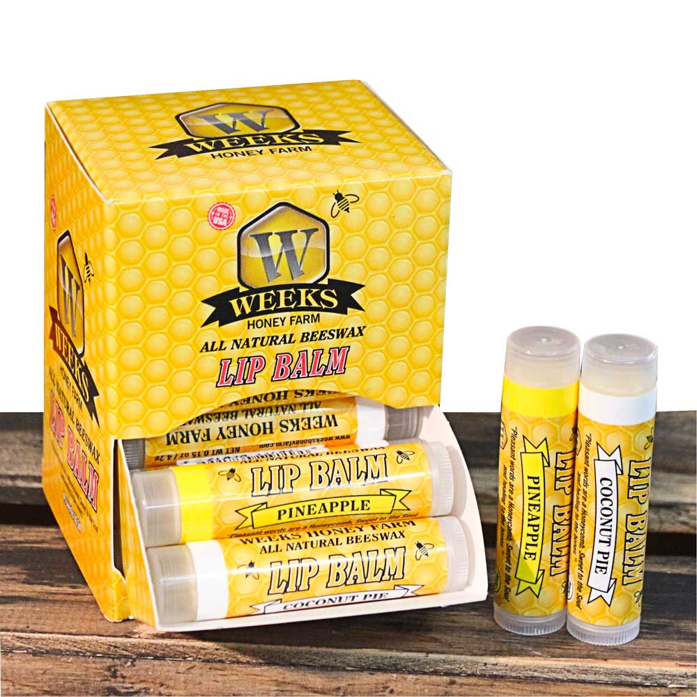 A Weeks Honey Farm display box labeled “ALL NATURAL BEESWAX LIP BALM” holds several Weeks All Natural Beeswax Lip Balms, including PINEAPPLE and COCONUT PIE flavors, arranged on a wooden surface. Singles, twin packs, and cases available.