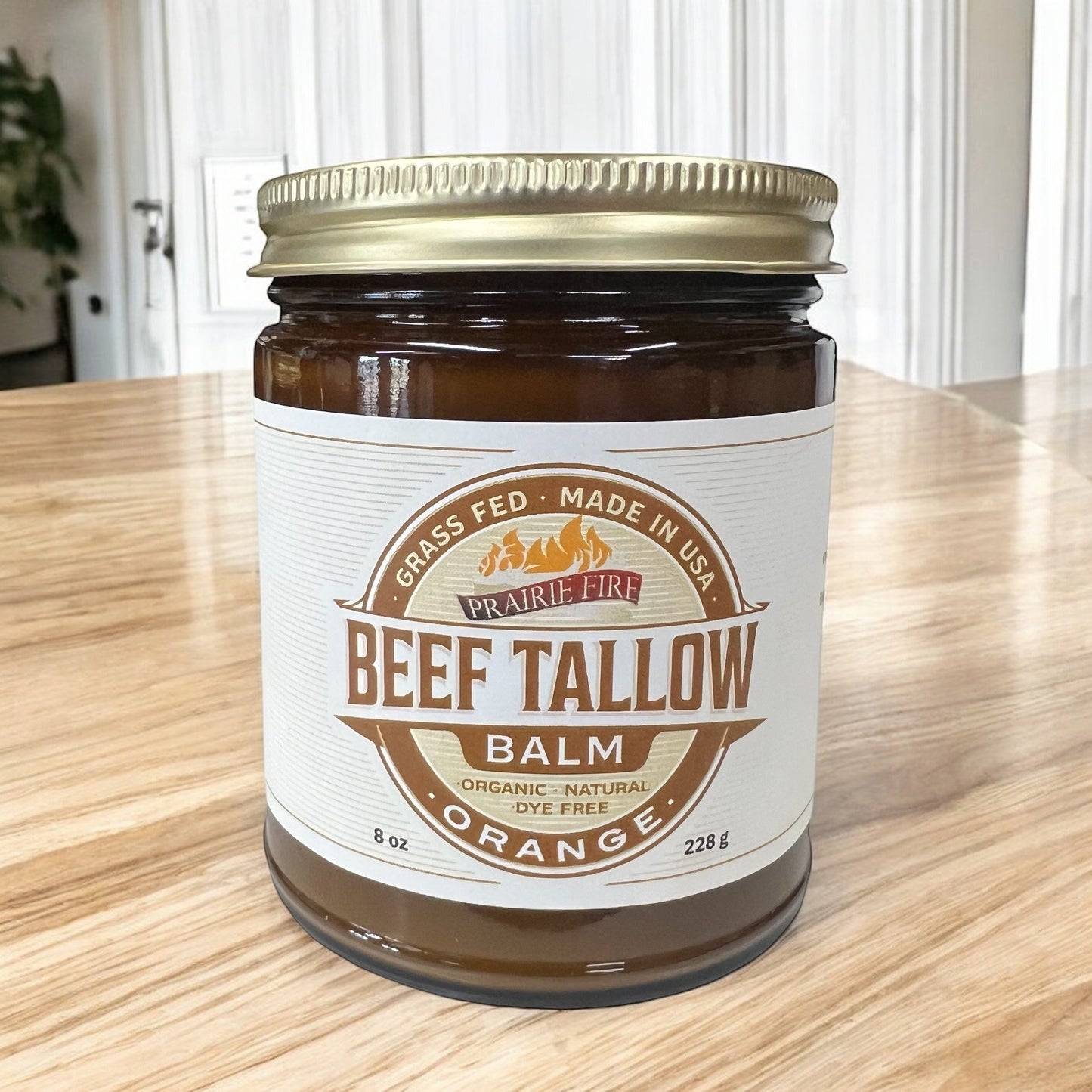 A brown glass jar of Prairie Fire Tallow, Candles, and Lavender’s Beef Tallow Balm (8 oz) with a gold lid sits on a wooden table. The organic, grass-fed balm is moisturizing, orange scented, dye-free, and made in the USA.