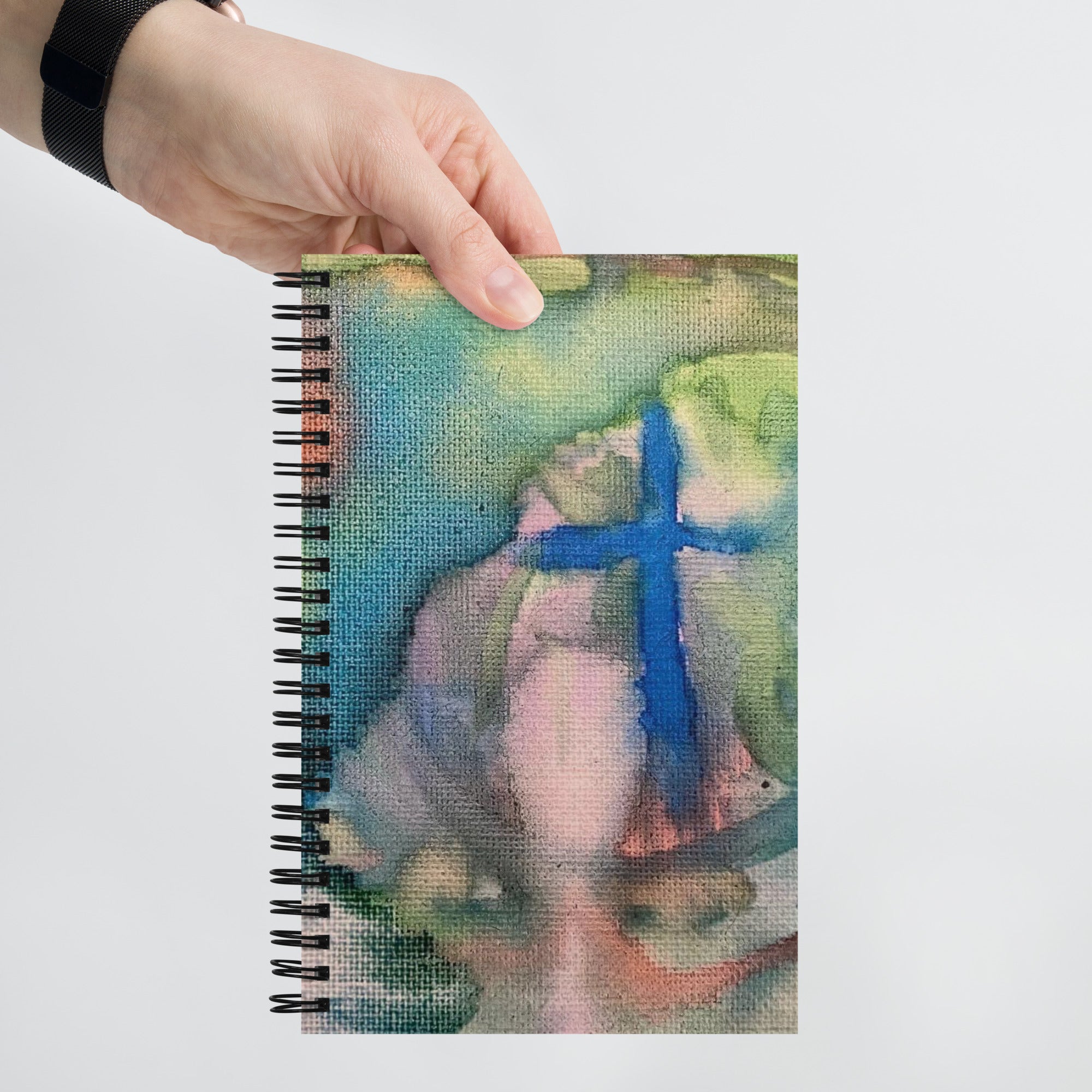 A hand holding a spiral-bound notebook with a colorful abstract cover featuring a prominent blue cross.