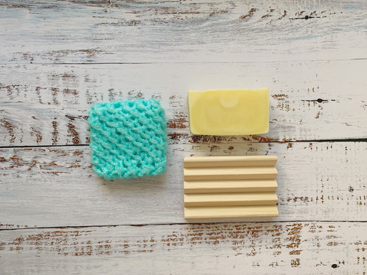 A light blue textured scrubber, a yellow bar of soap, and a beige Dot and Army Wooden Soap Dish (set of two) are arranged on a rustic white wooden surface.