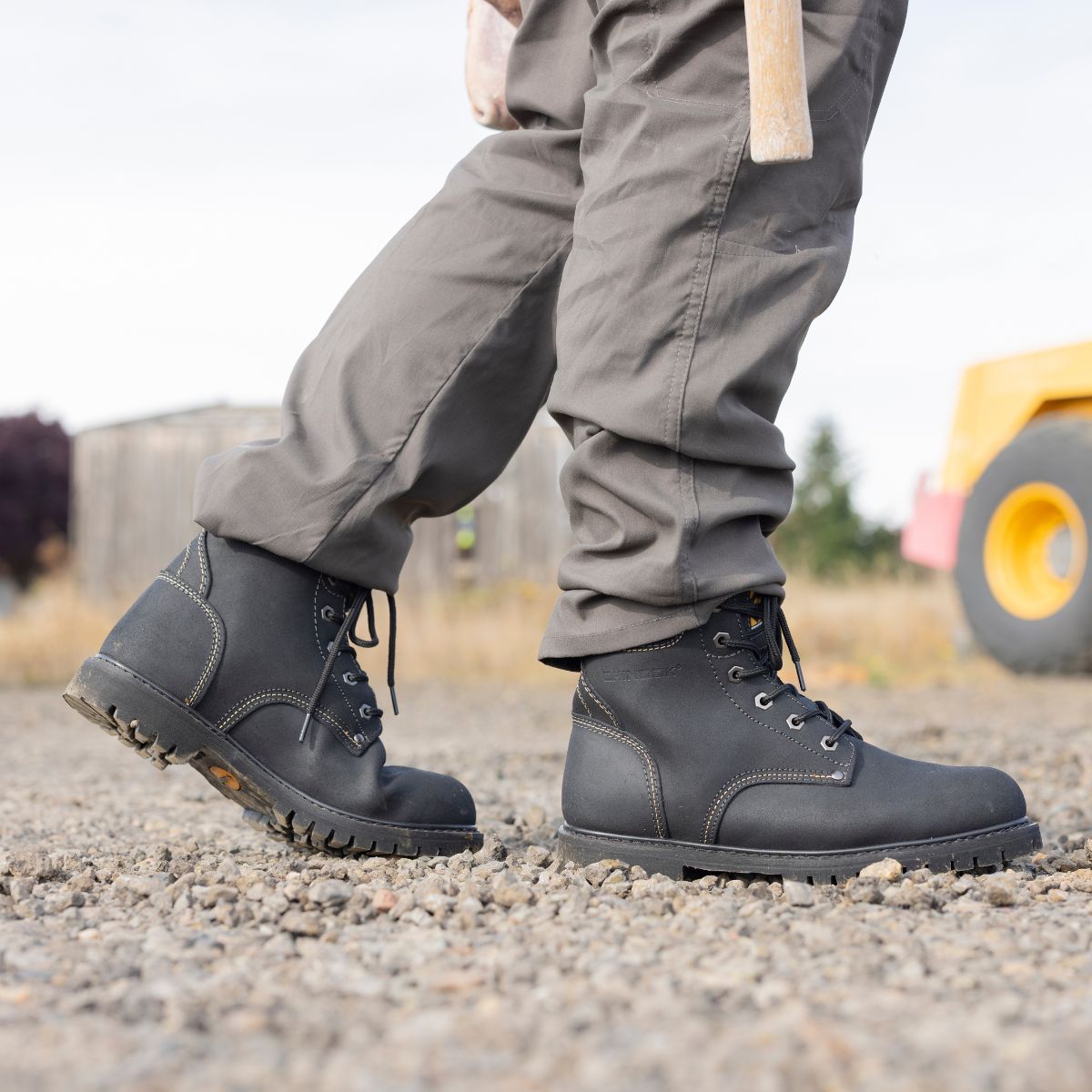 A person in gray work pants and Chinook® Footwear Oil Rigger 6 Steel Toe Men’s Work Boots (black) stands on gravel with an axe, while a yellow construction vehicle and wooden shed blur into the background, emphasizing rugged protection.