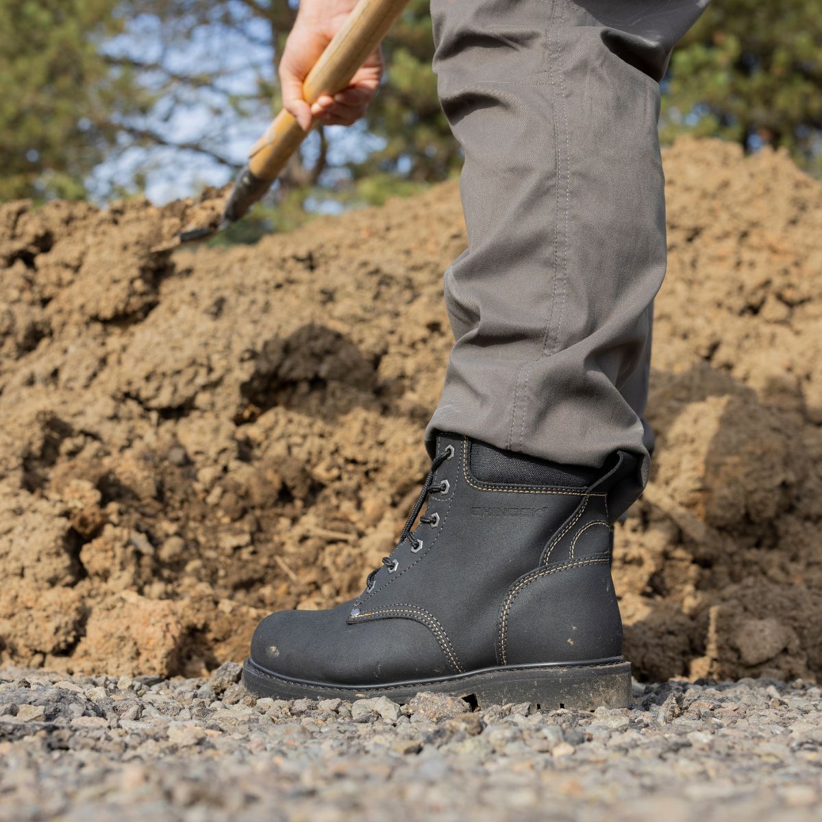 A person in gray pants and Chinook® Footwears Oil Rigger 6” Steel Toe Men’s Work Boots - Black stands on gravel with a shovel near a large soil pile, trees blurred in the background.